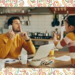 Father working from home on laptop and phone, gesturing for quiet as his child talks to him in the kitchen.